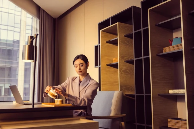 Young woman in blazer pours tea at modern desk with laptop, with contemporary bookshelf and city view through the window.