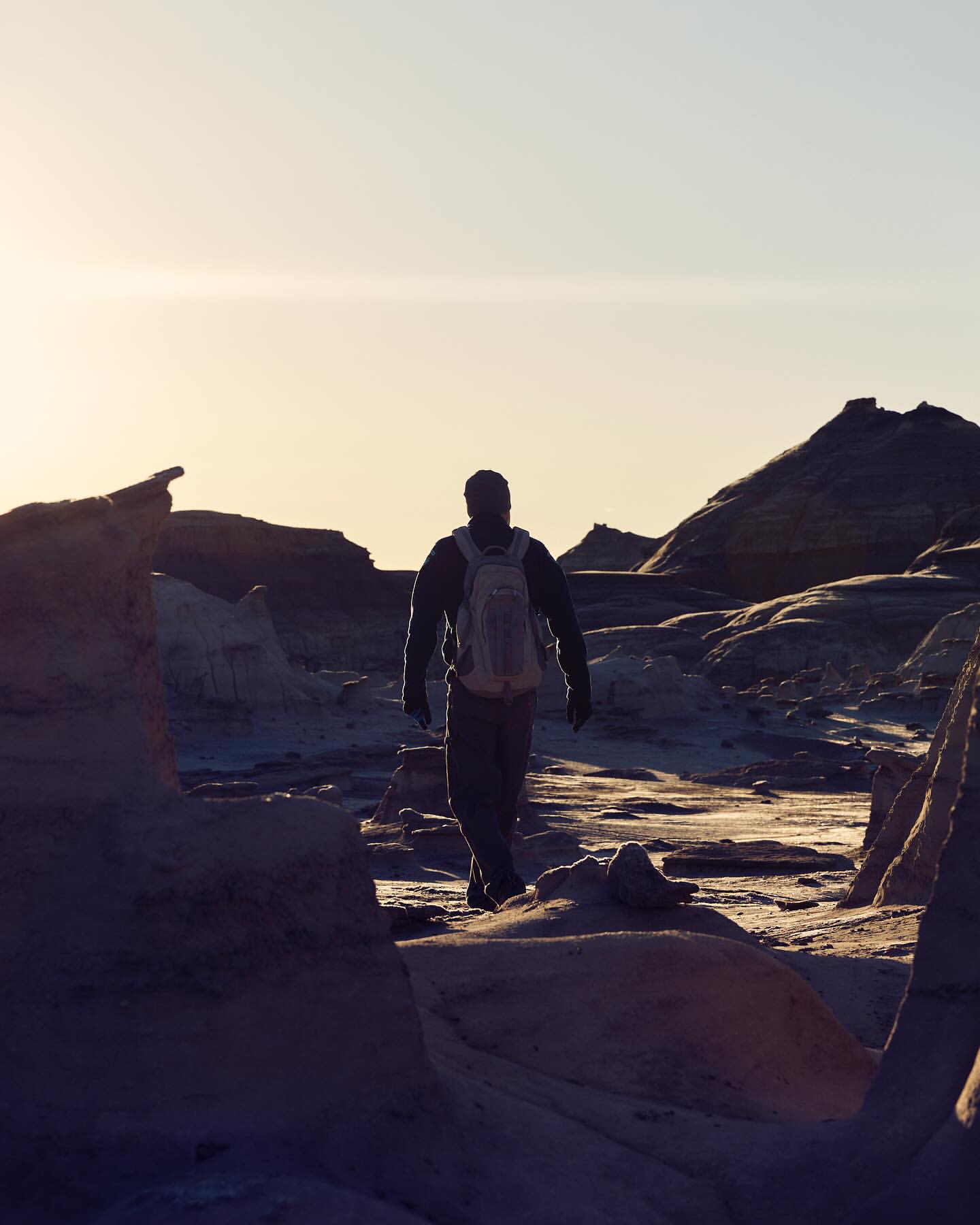 A person with a backpack walks away through a stark, otherworldly landscape under a bright sky, set against rock formations.