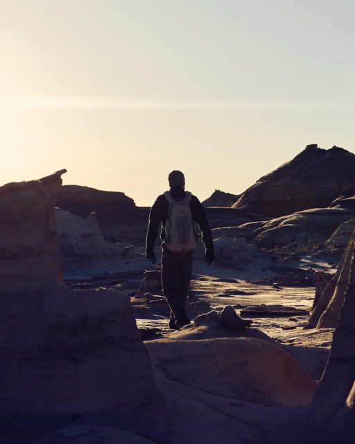 A person with a backpack walks away through a stark, otherworldly landscape under a bright sky, set against rock formations.