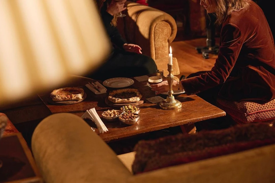 Two people enjoy an indoor gathering with pastries, nuts, and drinks arranged on a wooden table, illuminated by a candle.
