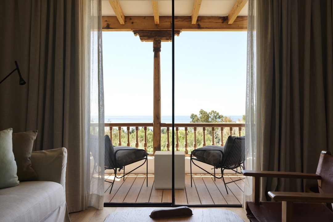 Interior view of a room with a balcony overlooking the sea. Wicker chairs and a white cube sit on the wooden balcony.