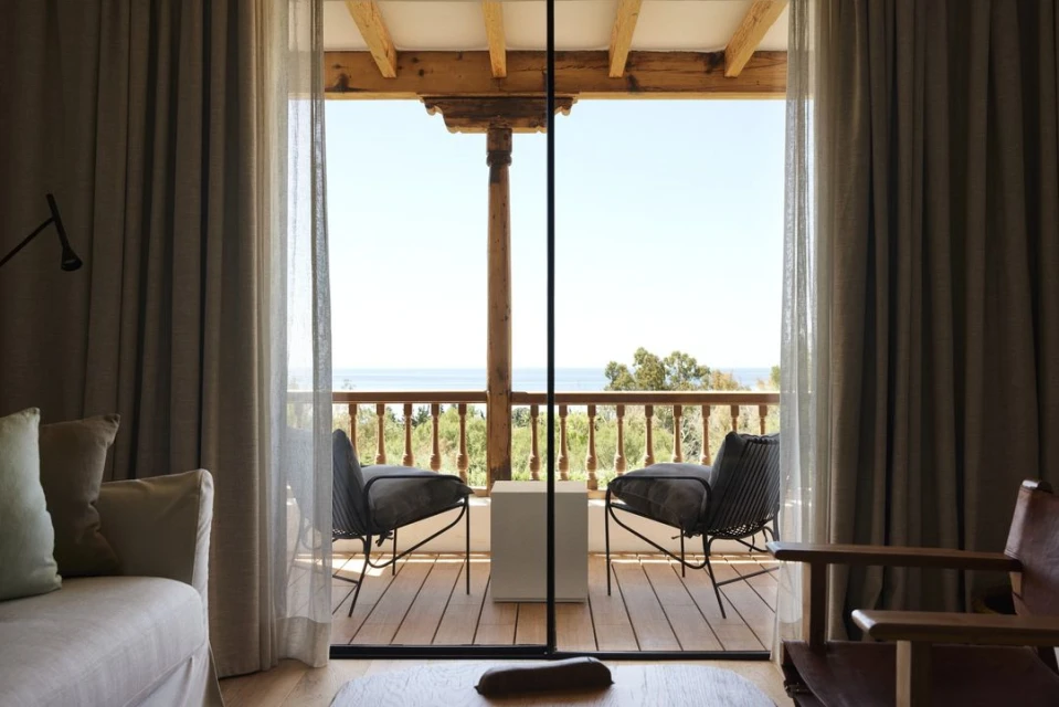 Interior view of a room with a balcony overlooking the sea. Wicker chairs and a white cube sit on the wooden balcony.