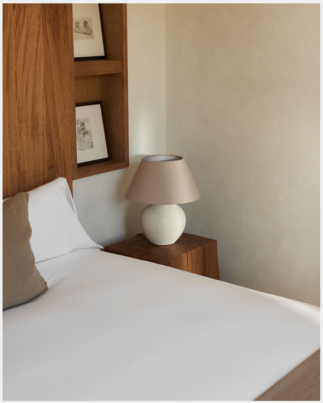 Bedroom interior featuring a bed with white sheets, a wooden side table, a table lamp, and framed artwork on the wooden headboard.