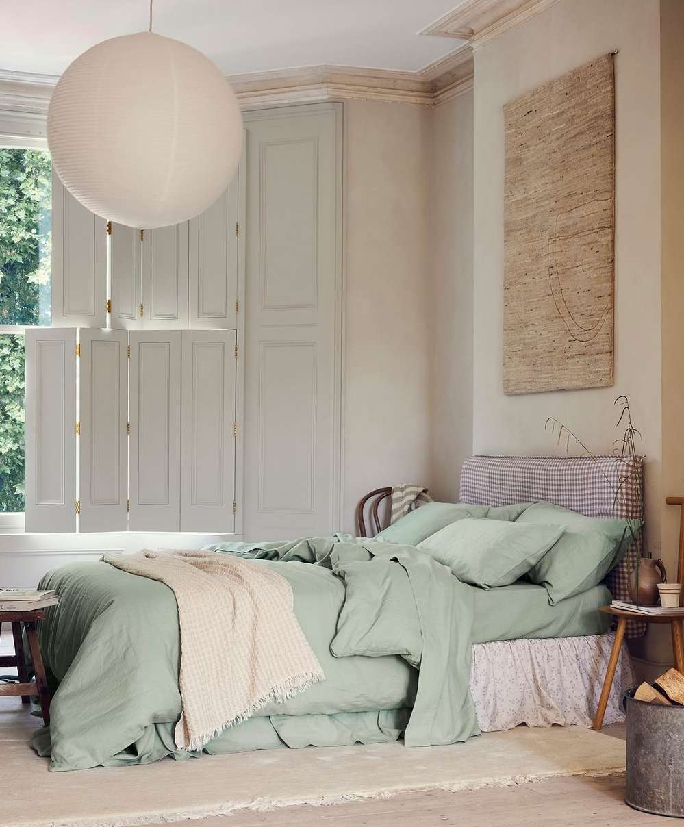 Interior of a bedroom featuring a bed with green bedding, a white blanket, wooden bedside tables and a large white round paper lamp.