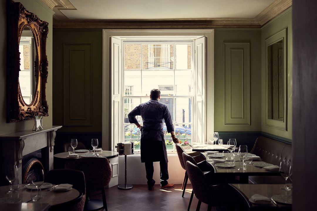 Adult male stands by window in a restaurant dining room; dining tables are set with wine glasses and plates in a sophisticated setting.