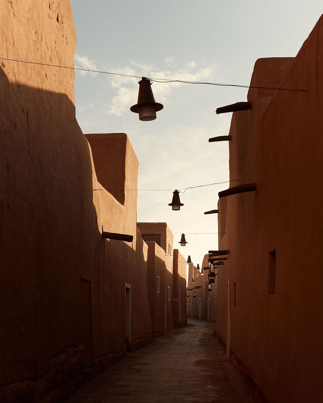 Narrow street lined with clay buildings and suspended lanterns, creating an atmospheric urban scene with soft lighting and architectural interest.
