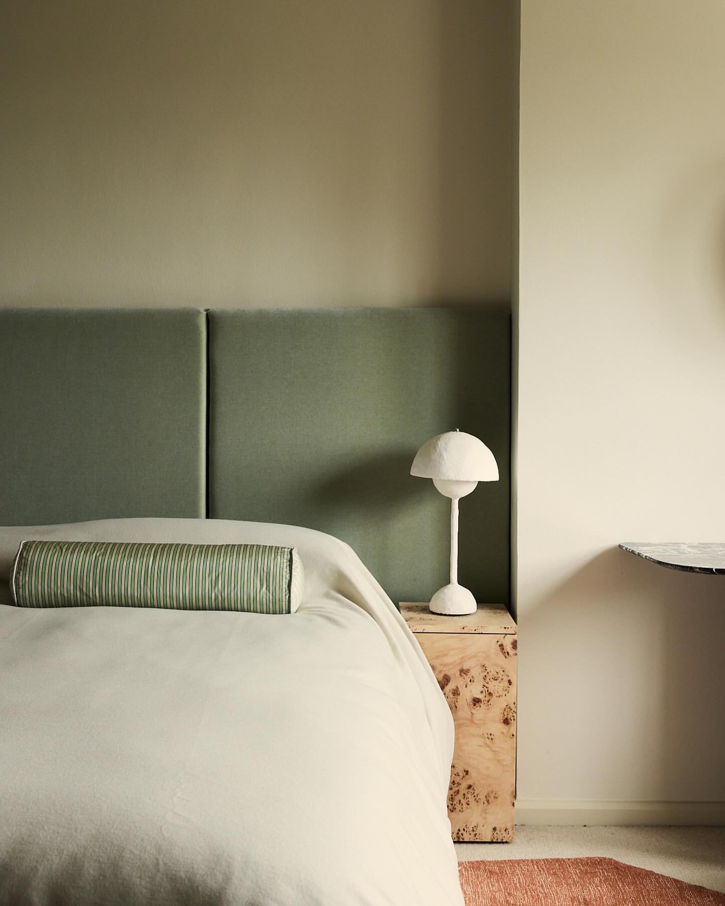 Bedroom interior with sage green headboard, bolster pillow, white lamp on a wooden nightstand. Light walls create a serene space.