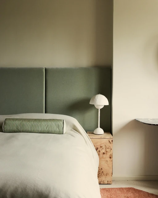 Bedroom interior with sage green headboard, bolster pillow, white lamp on a wooden nightstand. Light walls create a serene space.