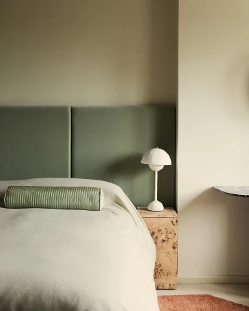 Bedroom interior with sage green headboard, bolster pillow, white lamp on a wooden nightstand. Light walls create a serene space.