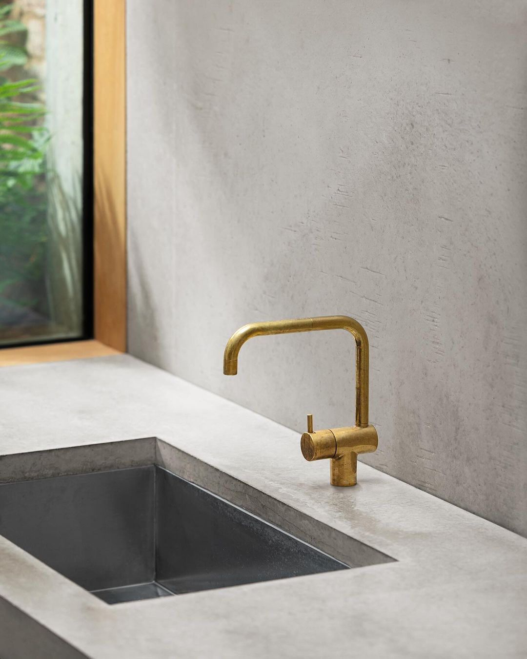 Modern bathroom interior with a rectangular gold faucet over a concrete sink, bathed in soft, natural light from a nearby window.