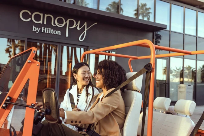 Two women smiling in an orange vehicle, with the Canopy by Hilton hotel in the background, captured in an eye-level outdoor shot.