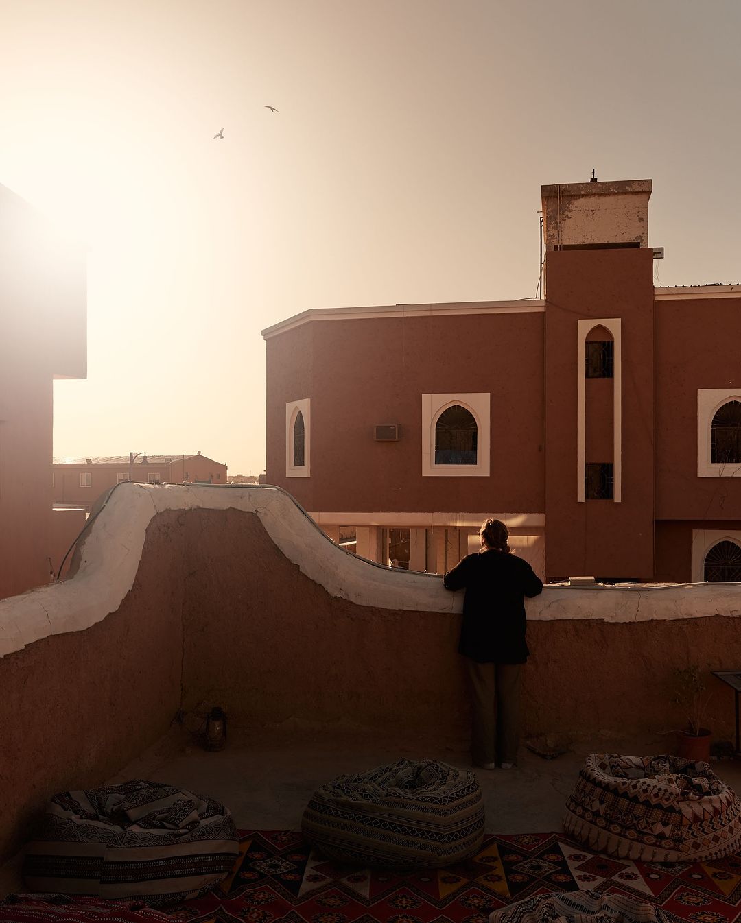 Adult looks over balcony with cityscape view and beanbag chairs in Morocco, bathed in the warm glow of setting sun, and blue sky.