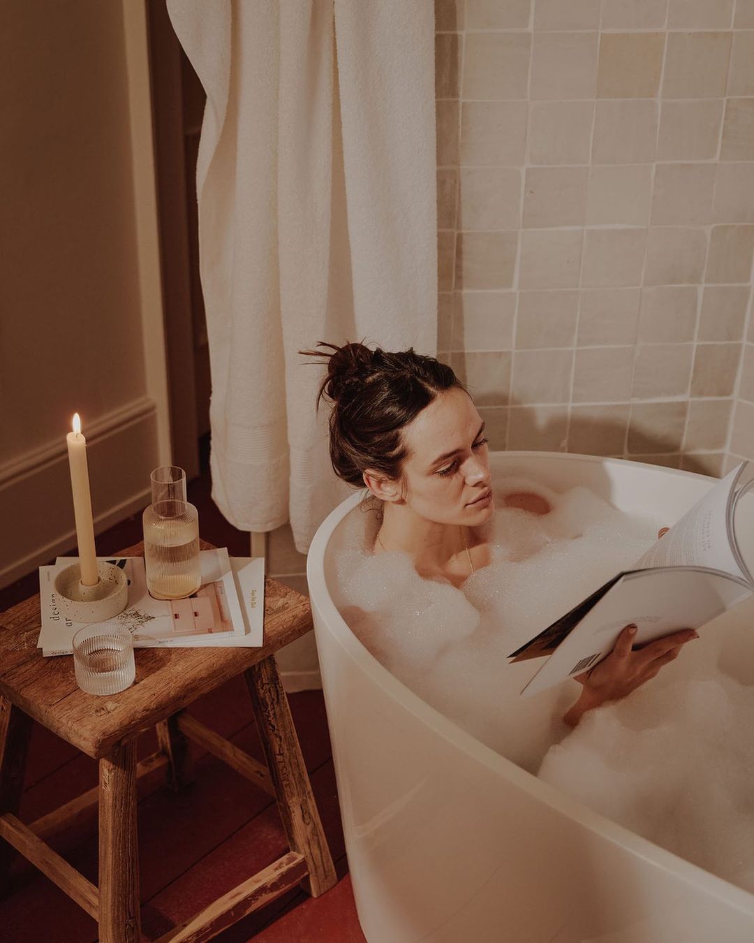 Woman relaxing in a bubble bath reading a magazine. Candle and glass of water are on wooden stool in a neutral-toned bathroom.