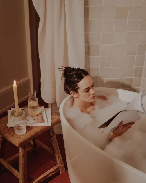 Woman relaxing in a bubble bath reading a magazine. Candle and glass of water are on wooden stool in a neutral-toned bathroom.