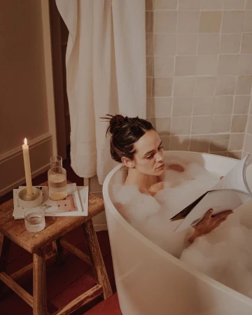 Woman relaxing in a bubble bath reading a magazine. Candle and glass of water are on wooden stool in a neutral-toned bathroom.