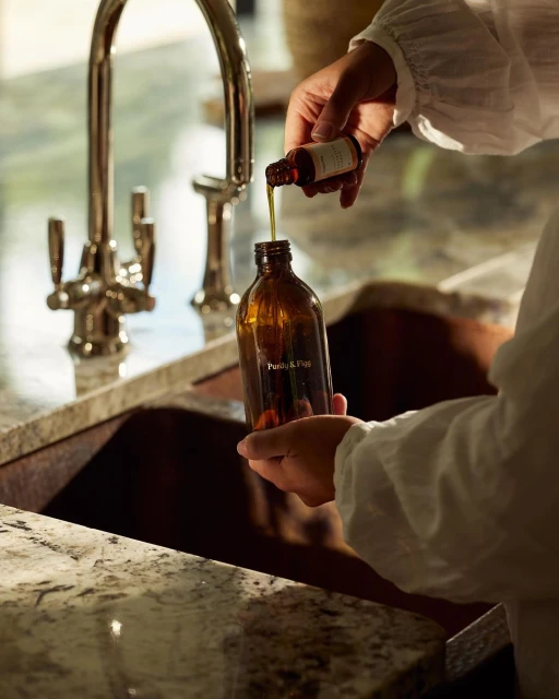 Person pours essential oil from small bottle into large amber glass bottle over granite countertop with metal faucet and copper sink.