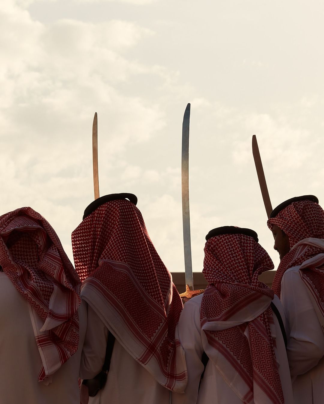 A group of men in traditional Arab clothing stand with swords raised against a neutral sky; likely a cultural event or celebration.