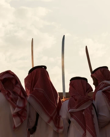 A group of men in traditional Arab clothing stand with swords raised against a neutral sky; likely a cultural event or celebration.