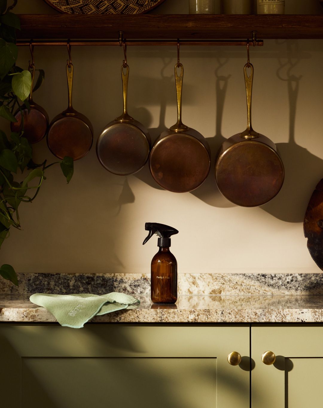 Kitchen still life of cleaning spray bottle and cloth on countertop with copper pots hanging on a rail against a light beige wall.