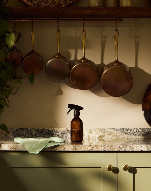 Kitchen still life of cleaning spray bottle and cloth on countertop with copper pots hanging on a rail against a light beige wall.