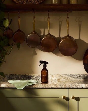 Kitchen still life of cleaning spray bottle and cloth on countertop with copper pots hanging on a rail against a light beige wall.