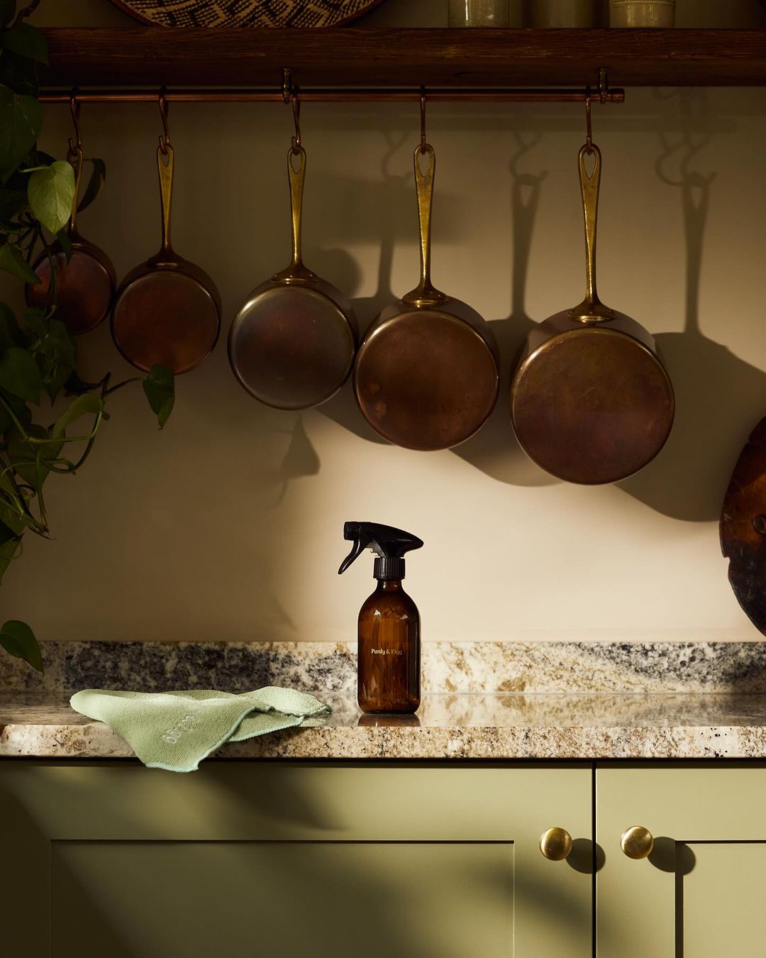 Still life shot of cleaning supplies on a kitchen counter with hanging pots and pans, featuring a brown spray bottle and green rag.