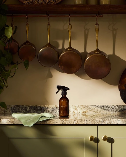 Still life shot of cleaning supplies on a kitchen counter with hanging pots and pans, featuring a brown spray bottle and green rag.
