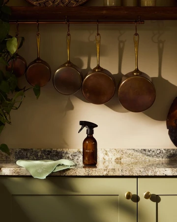 Still life shot of cleaning supplies on a kitchen counter with hanging pots and pans, featuring a brown spray bottle and green rag.