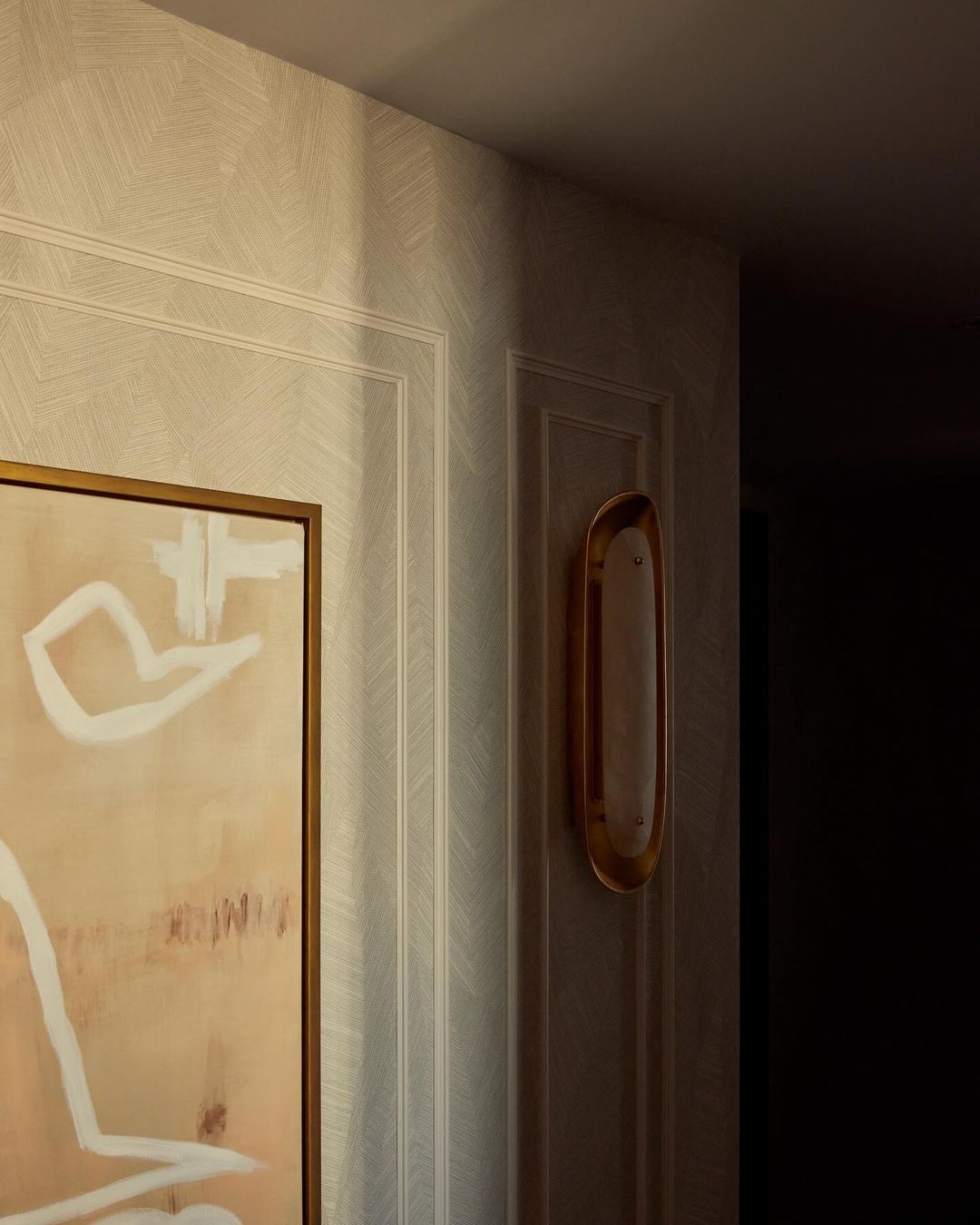 Interior design featuring an abstract painting with white and beige hues, complemented by a gold sconce on a textured wall.