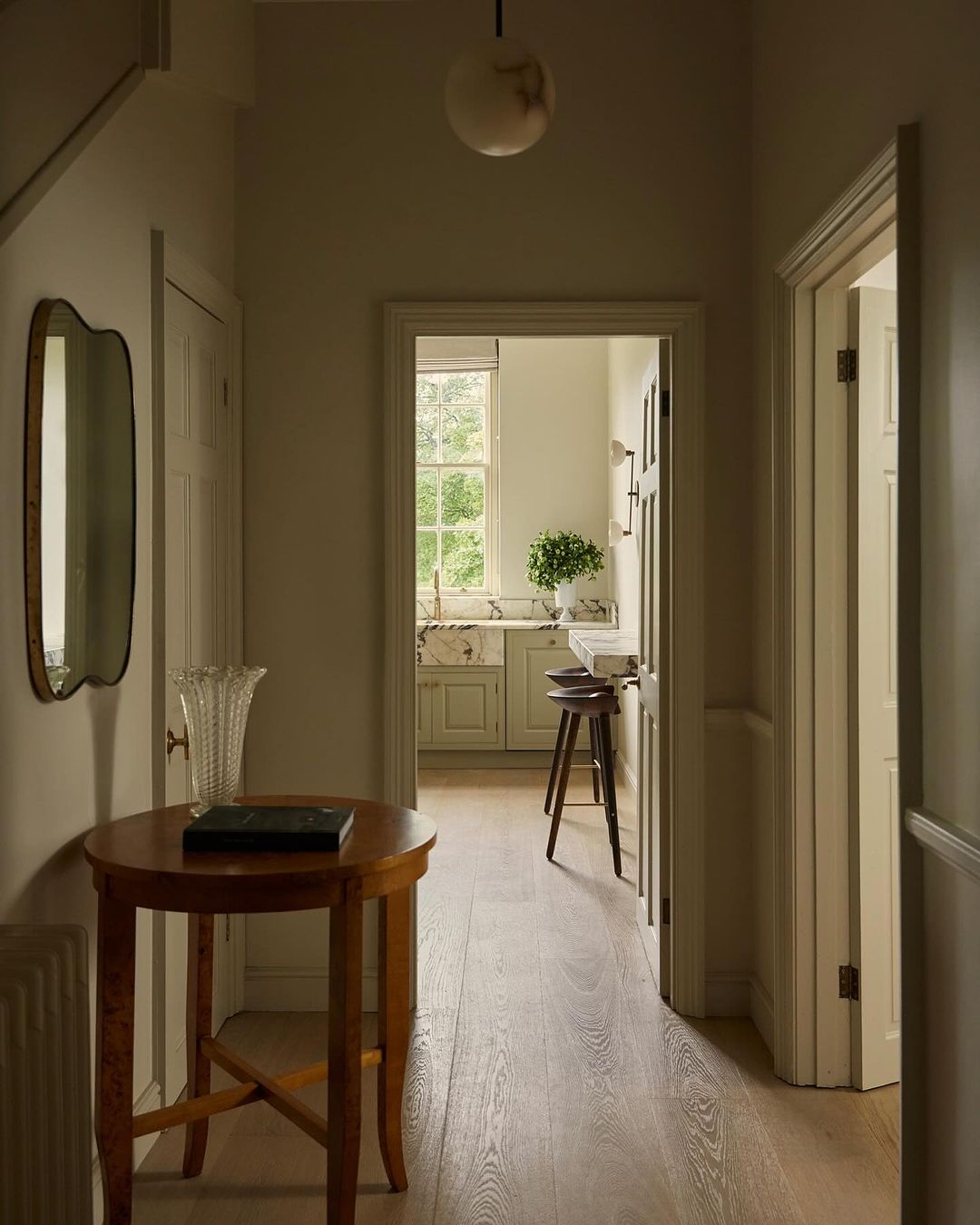 Interior view looking from hallway into kitchen, featuring wooden table, decorative vase, mirror, parquet floor, and neutral palette.