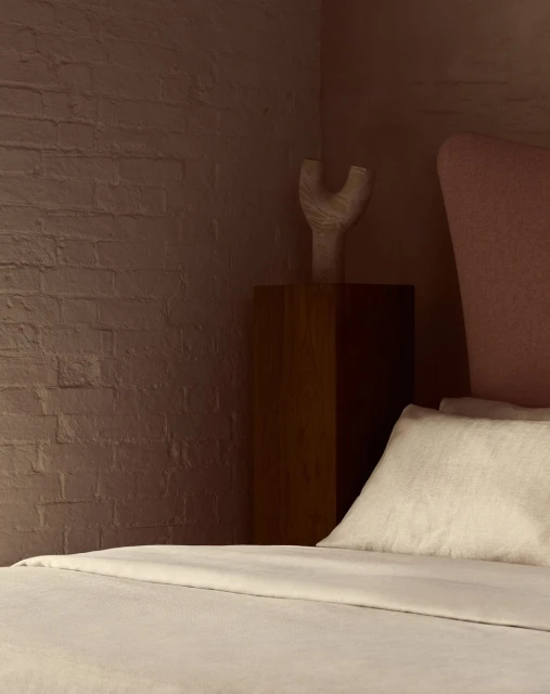 Bedroom interior with a cream-colored bed, light pink headboard, wooden nightstand, and a decorative white vase on top of the nightstand.