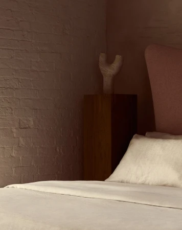 Bedroom interior with a cream-colored bed, light pink headboard, wooden nightstand, and a decorative white vase on top of the nightstand.