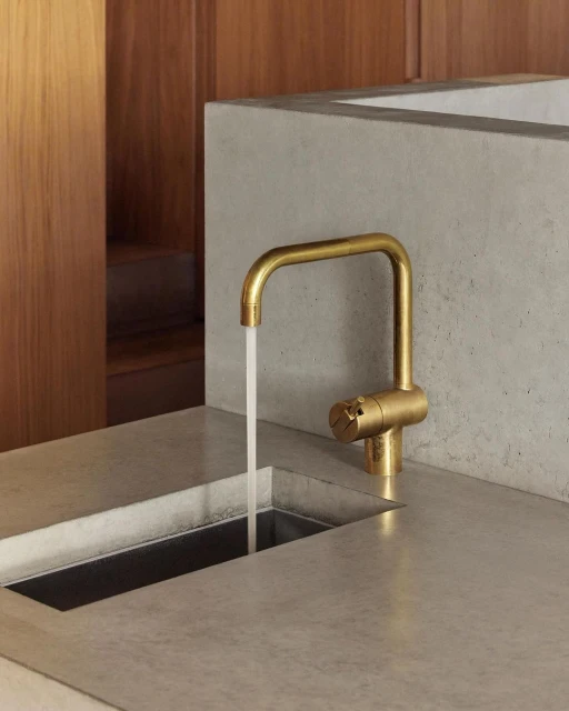 Close-up of a modern brushed gold faucet pouring water into a stainless steel sink set in a smooth, light grey countertop.