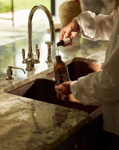 Person pours liquid from a small bottle into a brown glass bottle near a stainless steel kitchen faucet and granite countertop.