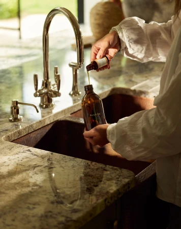 Person pours liquid from a small bottle into a brown glass bottle near a stainless steel kitchen faucet and granite countertop.