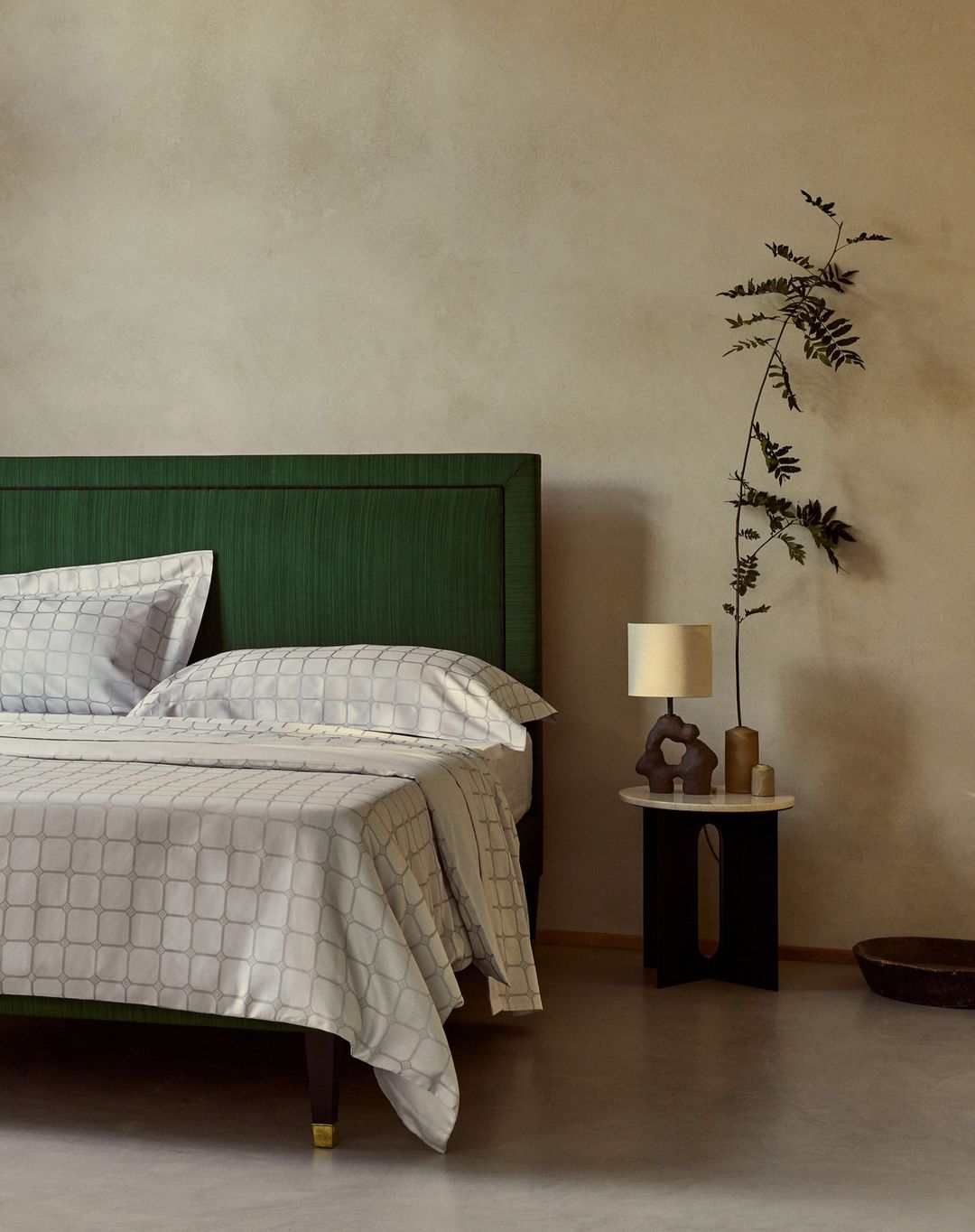 Bedroom interior featuring a bed with a green headboard and patterned linens, a small table, lamp, vase, and foliage against a neutral wall.
