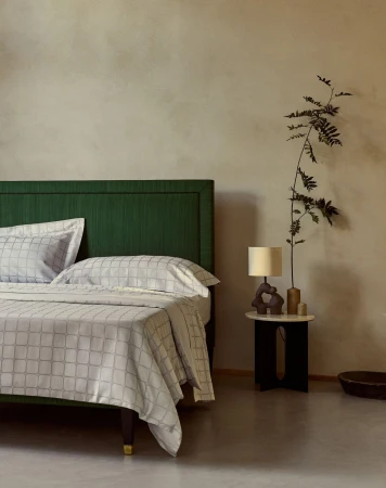 Bedroom interior featuring a bed with a green headboard and patterned linens, a small table, lamp, vase, and foliage against a neutral wall.