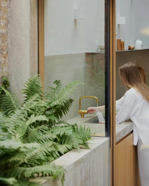 Woman in casual wear is standing near a sink in an indoor home setting, next to green ferns and pouring water from a golden faucet.
