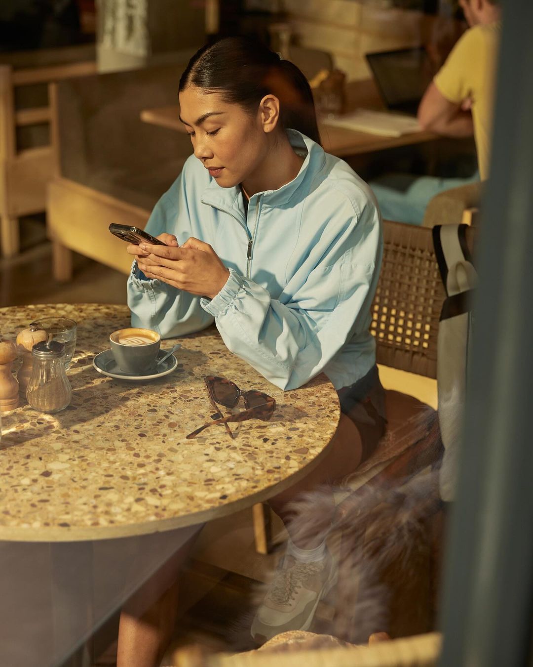 Woman in a blue jacket sits at a speckled stone table with coffee and sunglasses while using a phone in an indoor cafe.