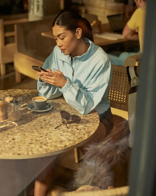 Woman in a blue jacket sits at a speckled stone table with coffee and sunglasses while using a phone in an indoor cafe.