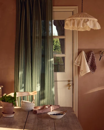 Indoor still life features a wooden table with dishware, linens, potted basil, and a view of greenery through a window with curtains.