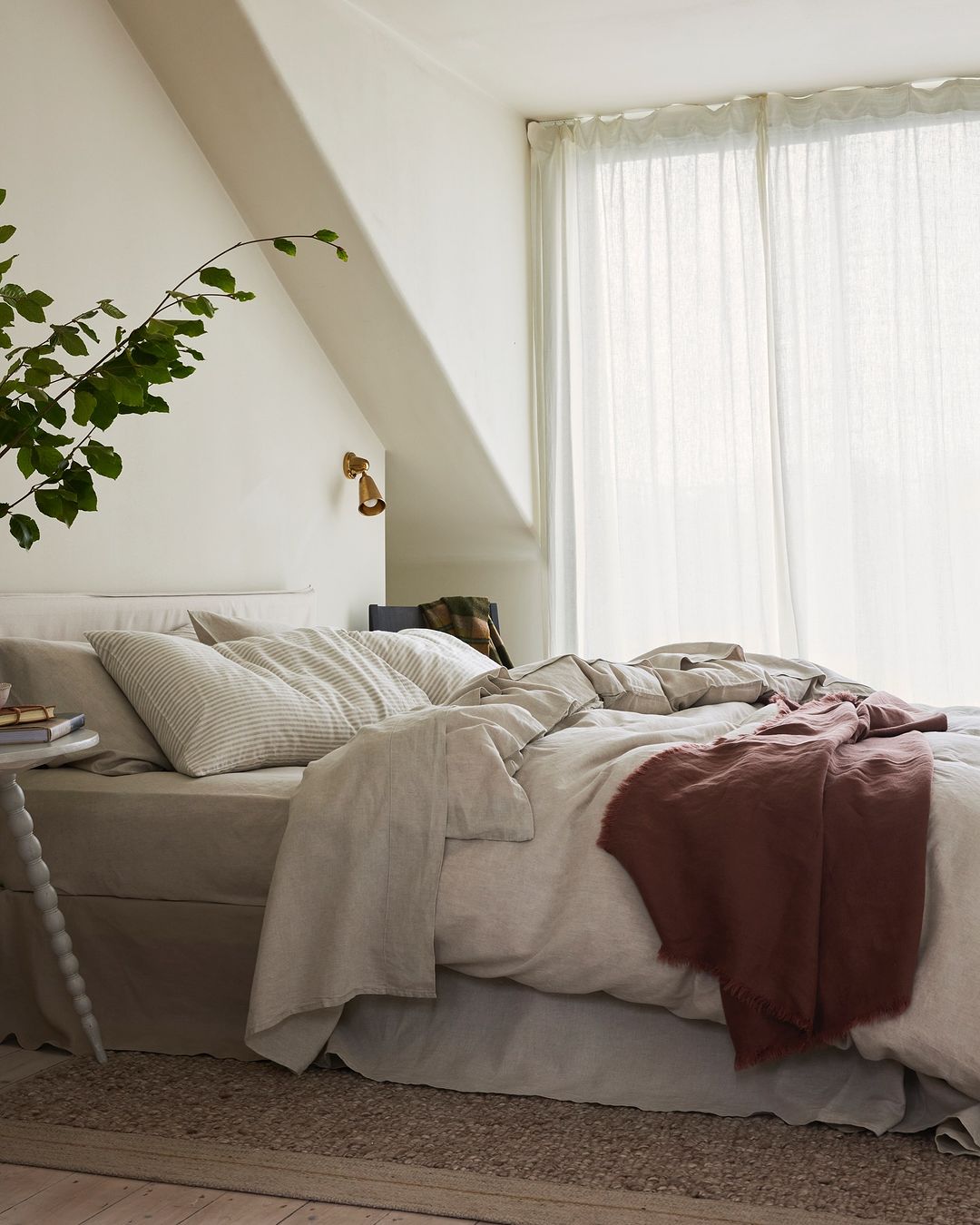 Bedroom interior featuring a neutral-toned bed with linen bedding, a small side table with books, and a sheer curtain in a minimalist style.