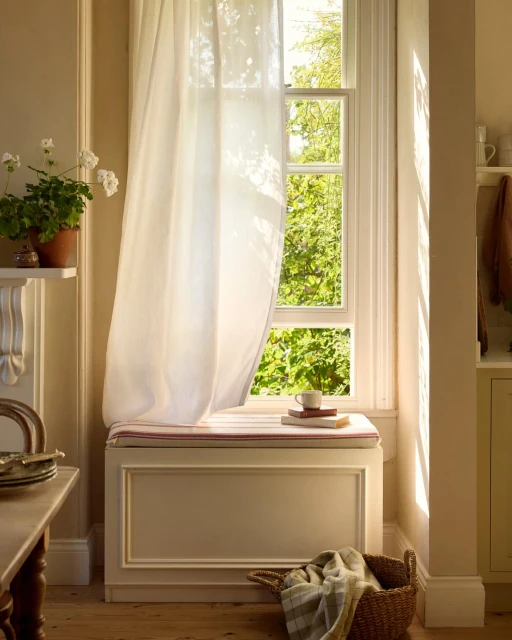 Bright, sunlit interior with a white-curtained window seat, books, a cup, white geraniums, wooden floors, and a woven basket with linen.