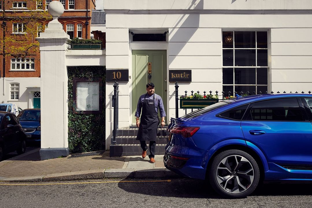 Blue Audi e-tron parked near a building with a man in an apron exiting the building's entrance, with warm light.