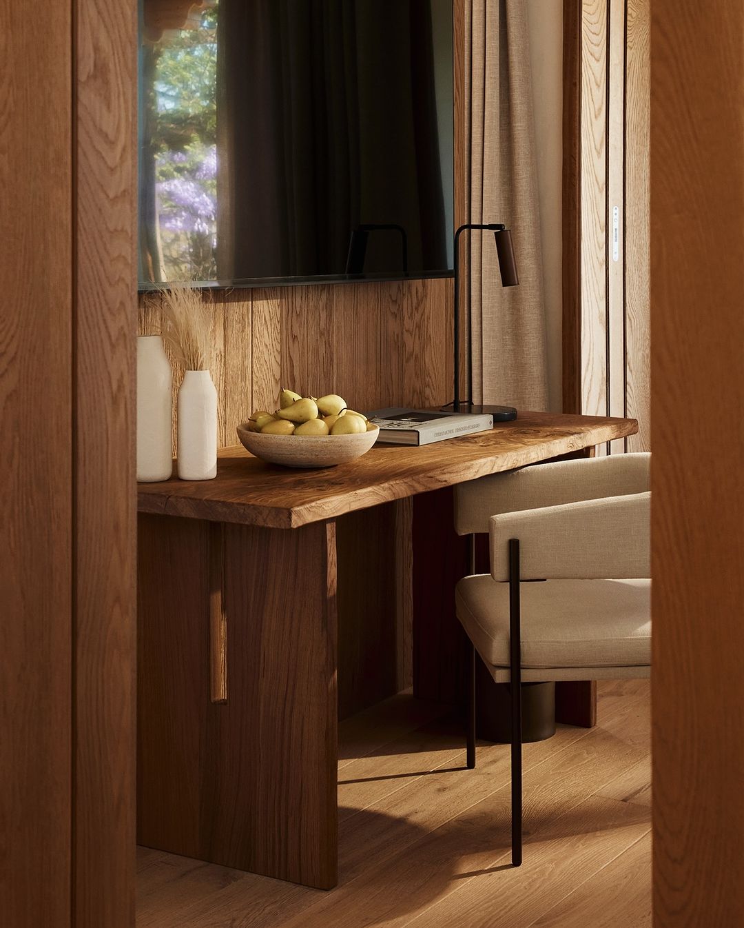 A desk setting with a wooden desk, a bowl of fruit, white vases, a modern chair, and a lamp in a well-lit interior.