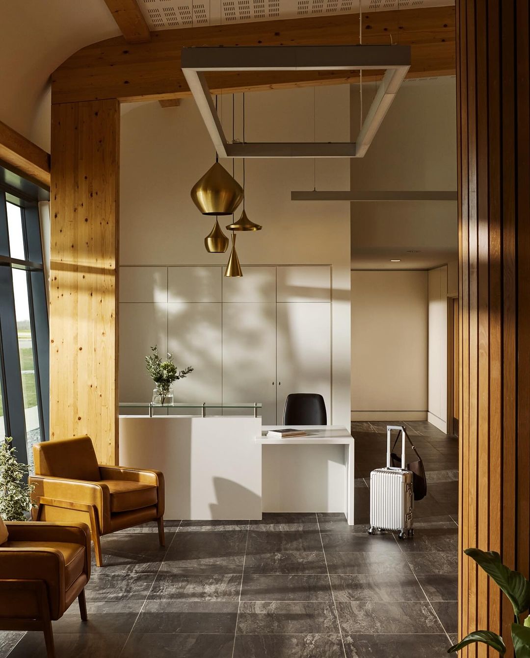 Lobby area with modern white reception desk, leather armchairs, suitcase, and contemporary lighting in an interior space.