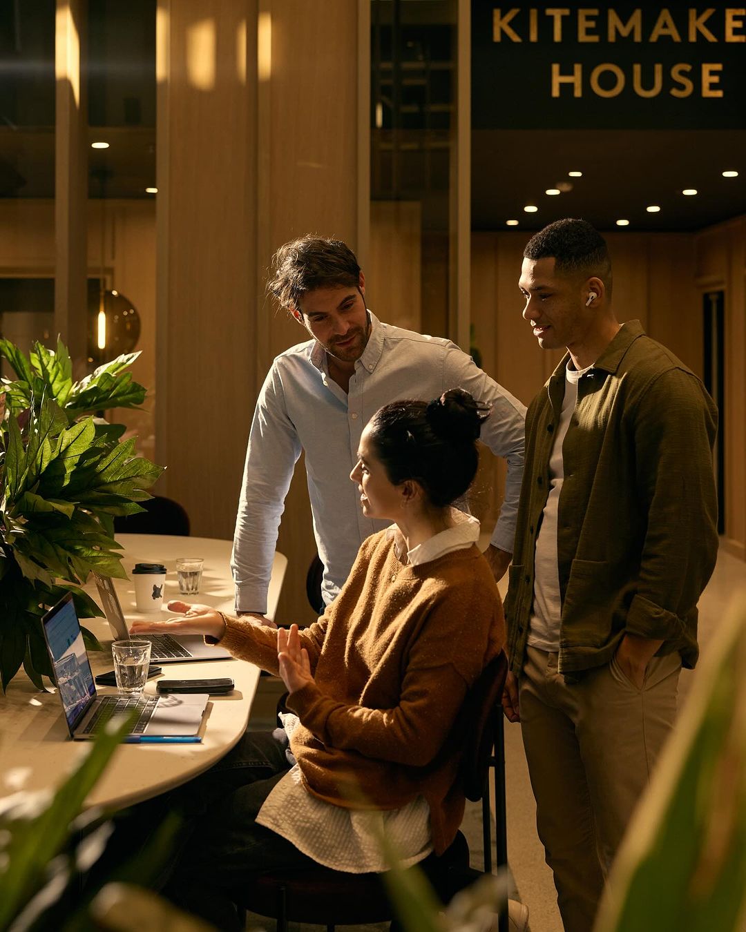Three colleagues gather around a table in a modern office, discussing a project on a laptop. Indoor setting with plants.