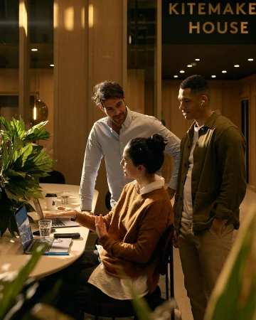 Three colleagues gather around a table in a modern office, discussing a project on a laptop. Indoor setting with plants.