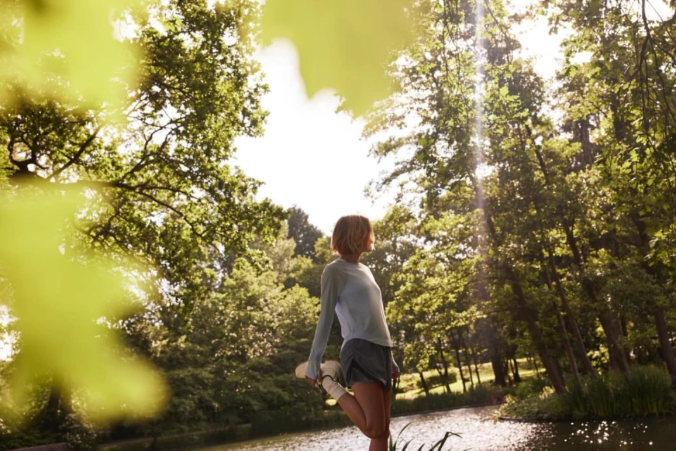 Woman stretching leg in sunlit forest setting. Green trees surround tranquil water with sky visible through foliage in this fitness shot.