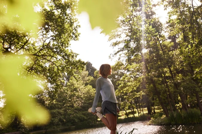 Woman stretching leg in sunlit forest setting. Green trees surround tranquil water with sky visible through foliage in this fitness shot.